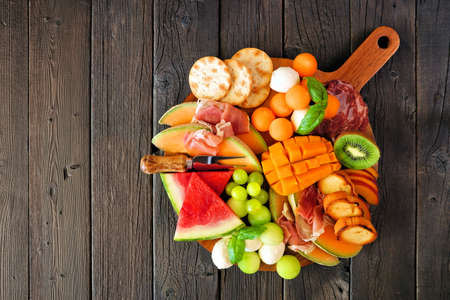Summer fruit theme charcuterie board against a dark wood background. Selection of fruits, cheese and meat appetizers. Top view.の写真素材