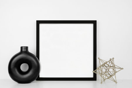 Mock up black square frame with modern home decor. White shelf against a white wall. Circle vase and...