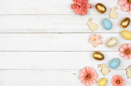Easter side border with eggs, paper flowers and wooden bunnies. Above view against a white wood...