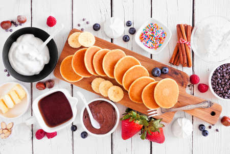 Breakfast or brunch pancake buffet table scene. Mini pancakes with a variety of toppings. Above view over a white wood background.の写真素材