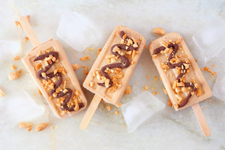 Peanut butter popsicles with chocolate drizzle. Top down view over a white marble background.