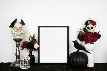 Halloween gothic romance mock up. Black frame on a black shelf with red and black flowers, skulls and pumpkin. Portrait frame against a white wall. Copy space.の写真素材