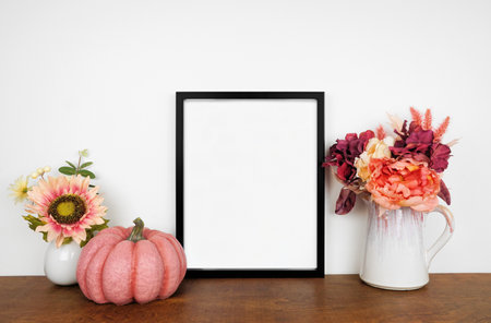 Mock up black picture frame with pink hue autumn flowers and pumpkin on a wood shelf against a white wall. Fall concept. Copy space.の写真素材