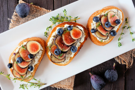 Crostini appetizers with figs, cream cheese spread and blueberries. Above view on a serving plate over a wood background.の写真素材