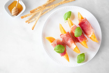 Fresh cantaloupe melon with prosciutto and basil. Above view table scene over a white marble background.の写真素材