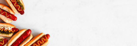 Variety of hot dogs with an assortment of toppings. Overhead view corner border on a white marble background.の写真素材