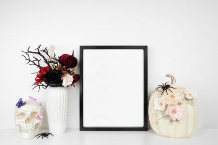 Halloween gothic romance mock up. Black frame on a white shelf with red and black flowers, skull and floral pumpkin. Portrait frame against a white wall. Copy space.の写真素材
