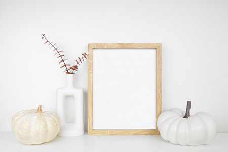 Mock up portrait wooden frame with fall braches and pumpkin decor on a white shelf against a white wall. Autumn concept. Copy space.の写真素材