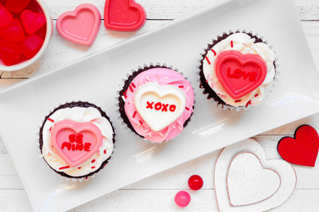 Fun Valentines Day cupcakes with messages on heart candies. Above view over a white wood background.