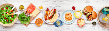 Summer BBQ or picnic table scene with hamburgers, hotdogs, salad and snacks. Above view over a white wood banner background.の写真素材