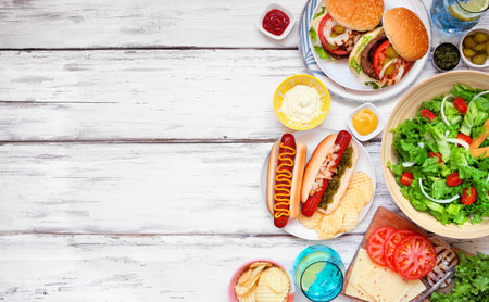 Summer BBQ or picnic side border with hamburgers, hotdogs, salad and snacks. Above view over a white wood background. Copy space.の写真素材