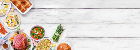 Traditional Easter ham dinner. Above view corner border on a white wood banner background with copy space. Ham, scalloped potatoes, vegetable side dishes, eggs, breads and lemon cake dessert.の写真素材