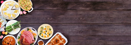 Classic Easter ham dinner. Overhead view corner border on a dark wood banner background with copy space. Ham, scalloped potatoes, vegetable side dishes, eggs, breads and lemon cake dessert.の写真素材