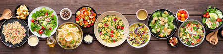 Assortment of delicious homemade salads. Overhead view table scene on a dark wood banner background. Summer food concept. Potato, caesar, pasta and green salads.の写真素材