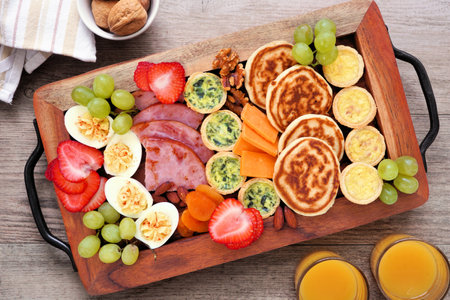 Breakfast or brunch grazing board on a wood tray. Variety of items including fruit, ham, quiche, pancakes and eggs. Top view over a wood table background.の写真素材