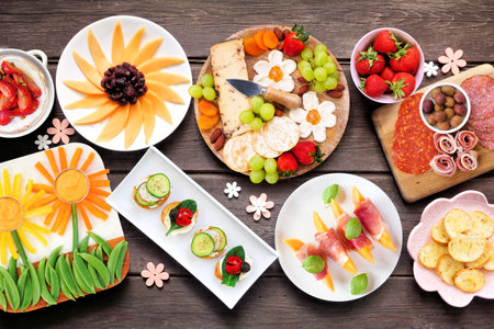 Summer garden party food table scene. Assortment of fun flower theme charcuterie items, cheese, fruit, vegetables and finger snacks. Top down view on a dark wood background.の写真素材