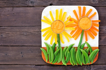 Fun summer theme vegetable snack board against a dark wood background. Flower arranged veggies with hummus. Above view.の写真素材