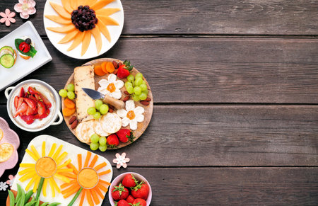 Summer garden party food side border. Variety of fun flower theme charcuterie items, cheese, fruit, vegetables and finger snacks. Top view on a dark wood background.の写真素材