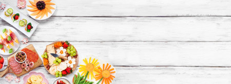Summer garden party food corner border. Assortment of fun flower theme charcuterie items, cheese, fruit, vegetables and finger snacks. Above view on a white wood banner background.の写真素材