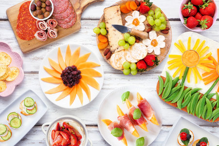 Summer garden party food table scene. Variety of fun flower theme charcuterie items, cheese, fruit, vegetables and finger snacks. Overhead view on a white wood background.の写真素材