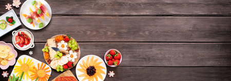 Summer garden party food corner border. Assortment of fun flower theme charcuterie items, cheese, fruit, vegetables and finger snacks. Above view on a dark wood banner background.の写真素材