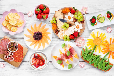 Summer garden party food table scene. Assortment of fun flower theme charcuterie items, cheese, fruit, vegetables and finger snacks. Top down view on a white wood background.の写真素材
