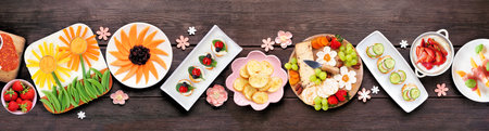 Summer garden party food table scene. Variety of fun flower theme charcuterie items, cheese, fruit, vegetables and finger snacks. Overhead view on a dark wood banner background.の写真素材