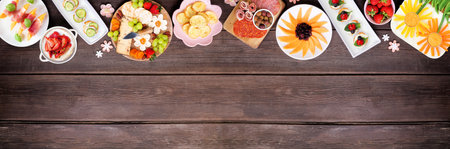 Summer garden party food top border. Assortment of fun flower theme charcuterie items, cheese, fruit, vegetables and finger snacks. Above view on a dark wood banner background.の写真素材