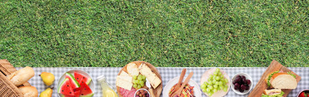 Summer picnic food bottom border. Top view with blanket over a green lawn banner background. Picnic basket, fruit, salad, sandwiches and snacks. Copy space.の写真素材