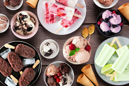 Cool summer food table scene. Assortment of ice cream bowls, popsicles and frozen treats. Above view on a dark wood background.の写真素材