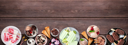 Cool summer food bottom border. Variety of ice cream bowls, popsicles and frozen treats. Top view on a dark wood banner background. Copy space.の写真素材