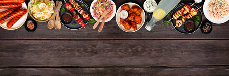 Summer BBQ food top border over a dark wood banner background. Variety of grilled meats, salads and sides. Overhead view.の写真素材