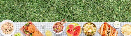 Summer picnic or BBQ food bottom border. Top down view with blanket over a green lawn banner background. Hamburgers, hot dogs, salads, fruit and lemonade. Copy space.の写真素材