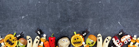 Healthy Halloween fruit and vegetable snacks. Variety of fun, spooky treats. Top down view bottom border over a dark stone banner background with copy space.の写真素材