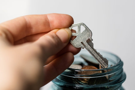 Hand holding a key in a glass jar with coins on white background