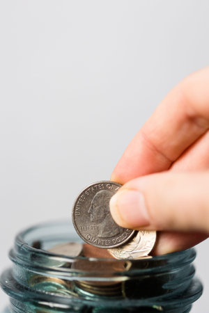 Close up of hand putting coins in a glass jar with white background, saving money concept