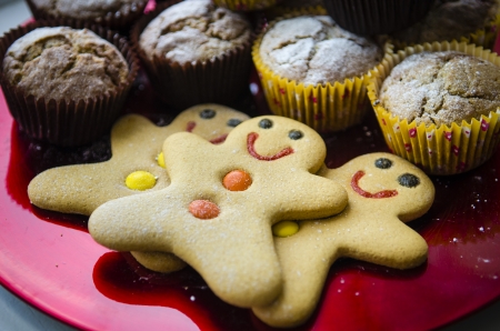 3 smiling happy gingerbread men with orange and yellow buttons on red Christmas metalic plate with muffins and dusted with icing sugar side viewの写真素材