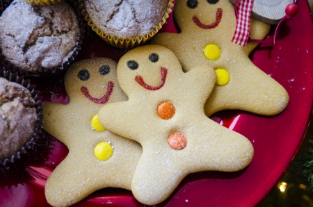 3 smiling happy gingerbread men with orange and yellow buttons on red Christmas metalic plate with muffins and dusted with icing sugarの写真素材
