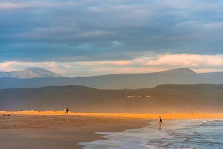 A streak of golden evening light falling on the Plettenberg Bay beach at sunset, with mountains in the distance and people walking on the beach. Garden Route, Western Cape, South Africaの写真素材