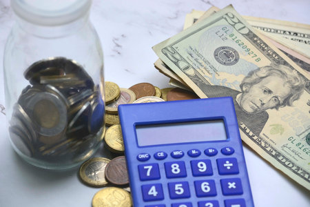 Glass jar with multicurrency coins, US dollar bills and calculator white table. Saving and...