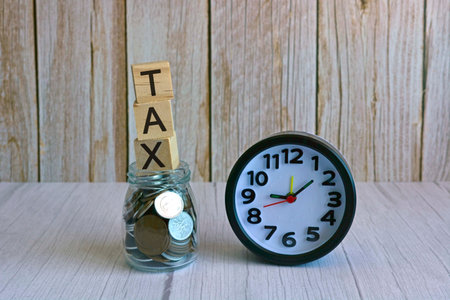 Tax text on wooden block cube on top of glass jar with multicurrency coins and alarm clock. Tax time conceptの写真素材
