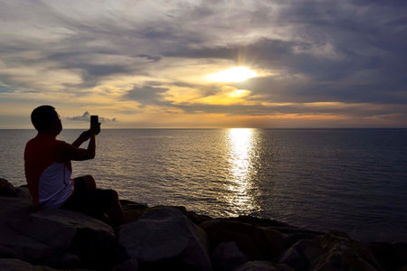 A man wearing face mask uses a smartphone to take pictures of beautiful sunset in Tanjung Aru Kota...