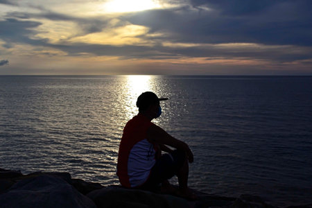 Man wearing facemask sitting on a rock watching the sunset at sea in Tanjung Aru Kota Kinabalu beach...