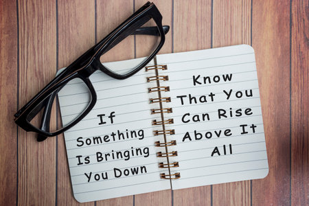 Motivational and inspirational quote on a notepad with black reading glass on wooden desk - If something is bringing you down, know what you can rise above it allの写真素材