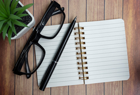 Notepad on wooden desk with potted plant, reading glasses and a pen. Directly above. Flat lay.