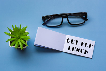 Out for lunch text on notepad with reading glasses and potted plant. Directly above. Flat lay,...