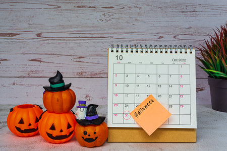 White October 2022 calendar with Jack o lantern pumpkin and potted plant on wooden desk. Halloween text on notepad.の写真素材