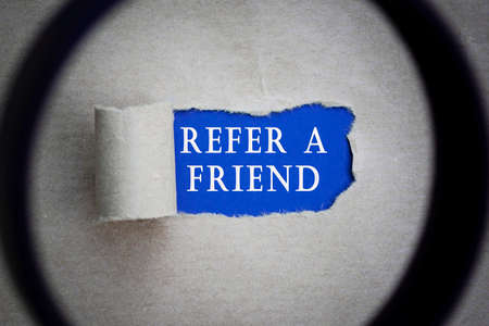 Refer A Friend text on torn hole in the sheet of brown paper on blue background with magnifying glass. Website, search and network concept.の写真素材