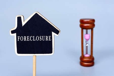 Foreclosure text on wooden house model with blurred hourglass background. Business, finance,...