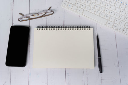 Notepad with computer keyboard, smartphone, pen and reading glasses on white wooden desk. Copy...